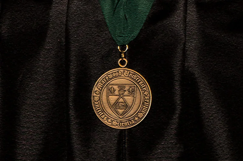Gold Medal with Marywood seal hanging from a green ribbon against black academic attire Marywood University to Award 39 Endowed Medals of Recognition to Graduates
