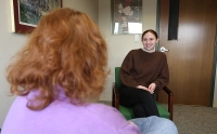 woman facing camera, sitting in green chair wearing brown sweater smiling and talking to woman facing away from camera with red hair and purple sweater