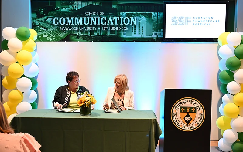 two women signing a document against a backlit wall and green, yellow, and white balloon decorations with a wall banner that says School of Communication and Scranton Shakespeare Festival Marywood University and Scranton Shakespeare Festival Announce Partnership