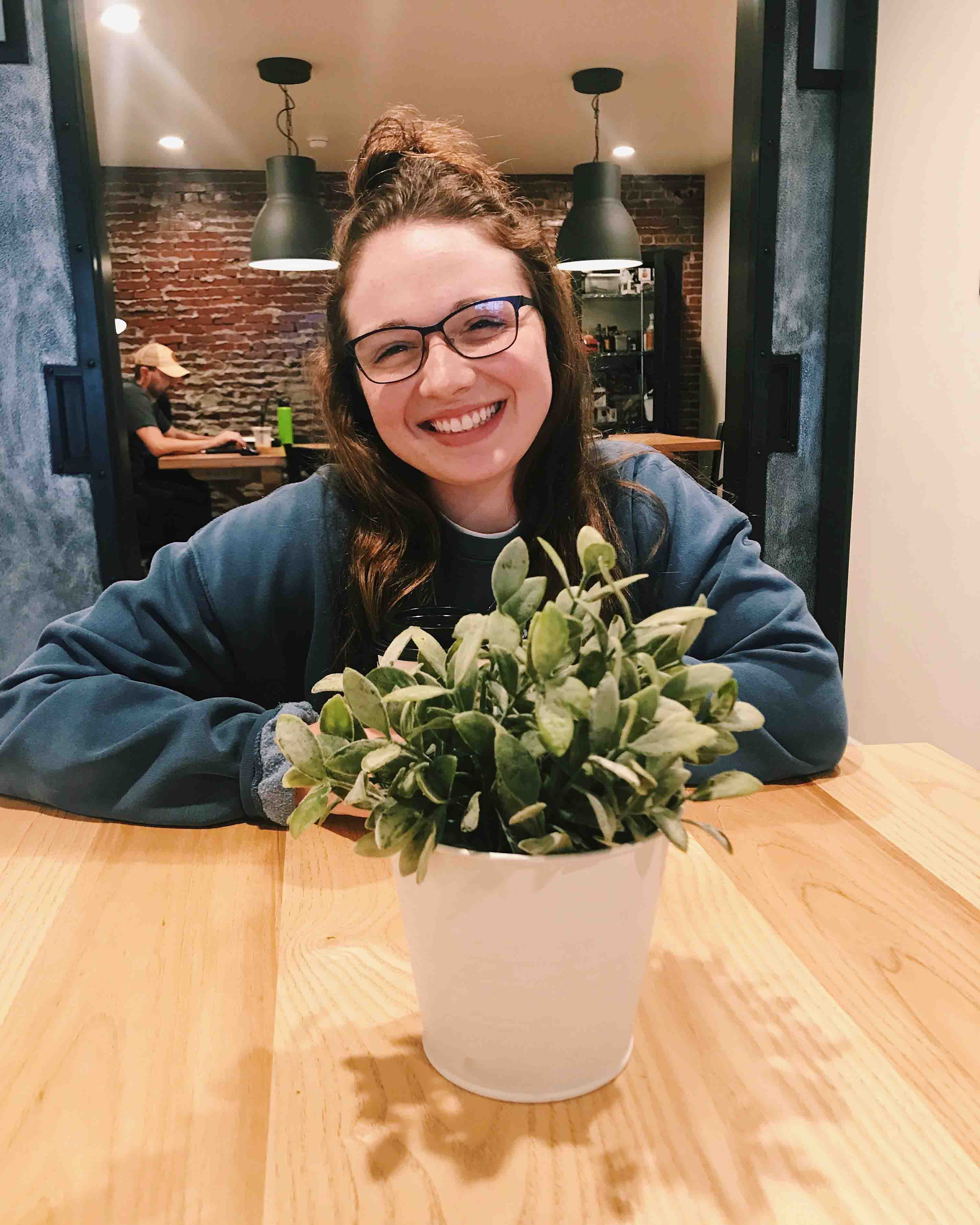 A young lady in glasses sitting in front of a potted plant.